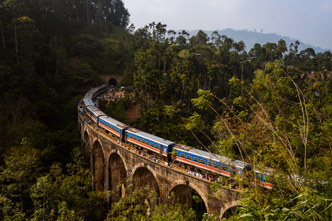 A train passes over a massive stone bridge that has nine arches and crosses a green jungle